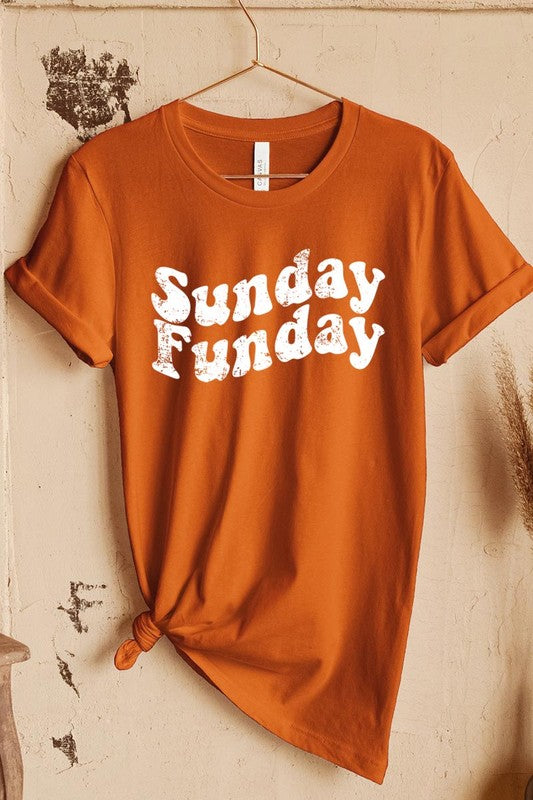 The "Sunday Funday" graphic tee with high-quality direct-to-film printing is paired with light blue distressed denim shorts, a brown hat, and a woven bag on a wooden hanger rack.