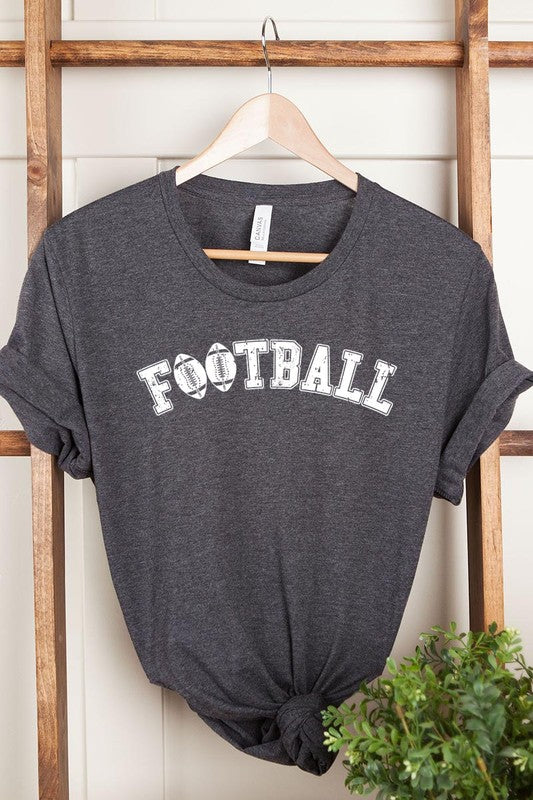 The Football Graphic Tee in beige features "FOOTBALL" in black lettering, paired with light blue distressed denim shorts and a pair of sunglasses. Crafted from premium materials for maximum comfort and style, this unisex crew neck short sleeve shirt is perfect for casual outings.