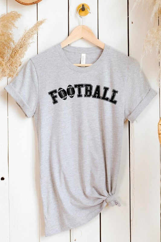 The Football Graphic Tee in beige features "FOOTBALL" in black lettering, paired with light blue distressed denim shorts and a pair of sunglasses. Crafted from premium materials for maximum comfort and style, this unisex crew neck short sleeve shirt is perfect for casual outings.