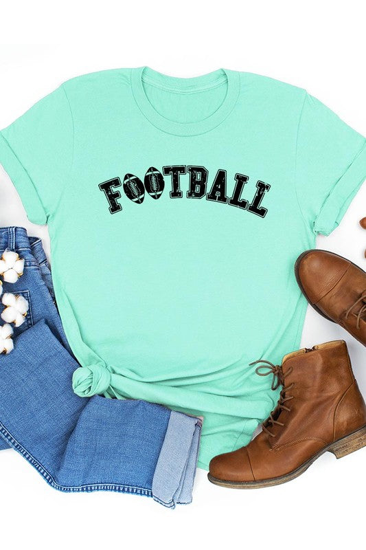 The Football Graphic Tee in beige features "FOOTBALL" in black lettering, paired with light blue distressed denim shorts and a pair of sunglasses. Crafted from premium materials for maximum comfort and style, this unisex crew neck short sleeve shirt is perfect for casual outings.