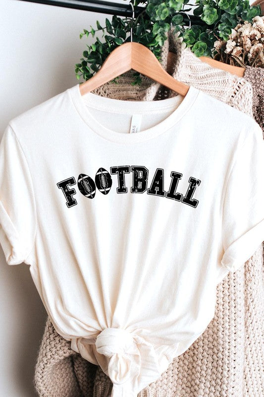 The Football Graphic Tee in beige features "FOOTBALL" in black lettering, paired with light blue distressed denim shorts and a pair of sunglasses. Crafted from premium materials for maximum comfort and style, this unisex crew neck short sleeve shirt is perfect for casual outings.