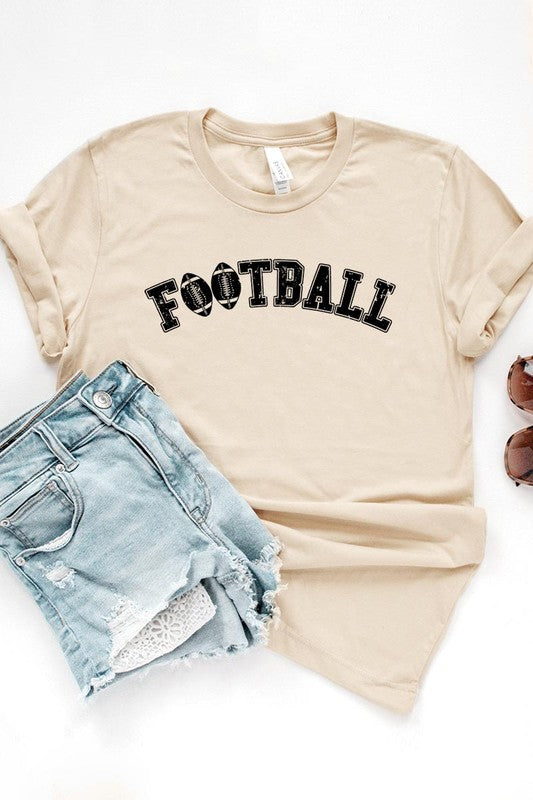 The Football Graphic Tee in beige features "FOOTBALL" in black lettering, paired with light blue distressed denim shorts and a pair of sunglasses. Crafted from premium materials for maximum comfort and style, this unisex crew neck short sleeve shirt is perfect for casual outings.