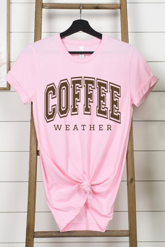 The Coffee Weather Graphic Tee, in a white hue with bold brown text, hangs on a wooden hanger, complemented by a beige sweater draped beside it and set against a background of lush green foliage. This unisex crew neck short sleeve tee showcases an eye-catching graphic design created through high-quality direct-to-film printing.