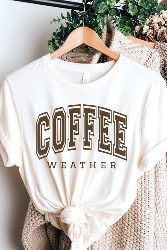 The Coffee Weather Graphic Tee, in a white hue with bold brown text, hangs on a wooden hanger, complemented by a beige sweater draped beside it and set against a background of lush green foliage. This unisex crew neck short sleeve tee showcases an eye-catching graphic design created through high-quality direct-to-film printing.