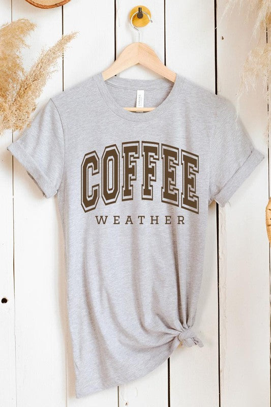 The Coffee Weather Graphic Tee, in a white hue with bold brown text, hangs on a wooden hanger, complemented by a beige sweater draped beside it and set against a background of lush green foliage. This unisex crew neck short sleeve tee showcases an eye-catching graphic design created through high-quality direct-to-film printing.