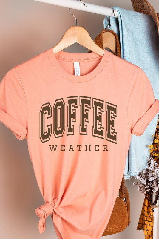 The Coffee Weather Graphic Tee, in a white hue with bold brown text, hangs on a wooden hanger, complemented by a beige sweater draped beside it and set against a background of lush green foliage. This unisex crew neck short sleeve tee showcases an eye-catching graphic design created through high-quality direct-to-film printing.