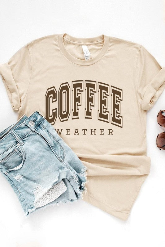 The Coffee Weather Graphic Tee, in a white hue with bold brown text, hangs on a wooden hanger, complemented by a beige sweater draped beside it and set against a background of lush green foliage. This unisex crew neck short sleeve tee showcases an eye-catching graphic design created through high-quality direct-to-film printing.