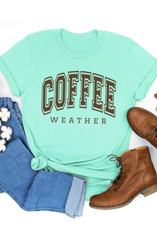 The Coffee Weather Graphic Tee, in a white hue with bold brown text, hangs on a wooden hanger, complemented by a beige sweater draped beside it and set against a background of lush green foliage. This unisex crew neck short sleeve tee showcases an eye-catching graphic design created through high-quality direct-to-film printing.