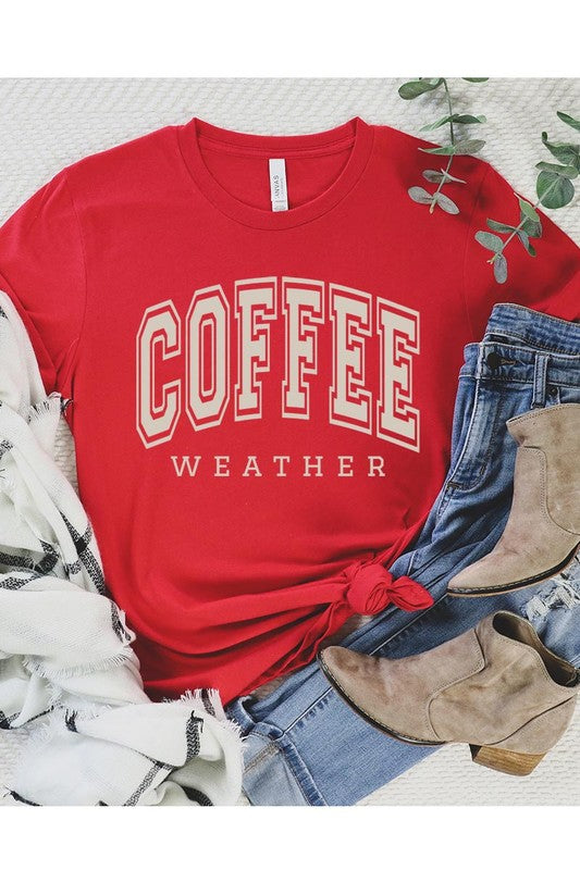 The Coffee Weather Graphic Tee, in a white hue with bold brown text, hangs on a wooden hanger, complemented by a beige sweater draped beside it and set against a background of lush green foliage. This unisex crew neck short sleeve tee showcases an eye-catching graphic design created through high-quality direct-to-film printing.