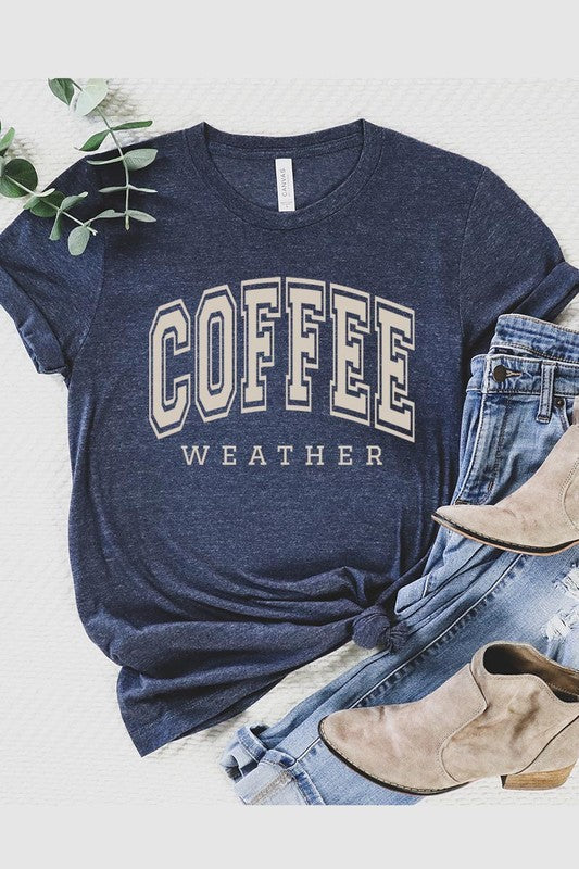 The Coffee Weather Graphic Tee, in a white hue with bold brown text, hangs on a wooden hanger, complemented by a beige sweater draped beside it and set against a background of lush green foliage. This unisex crew neck short sleeve tee showcases an eye-catching graphic design created through high-quality direct-to-film printing.