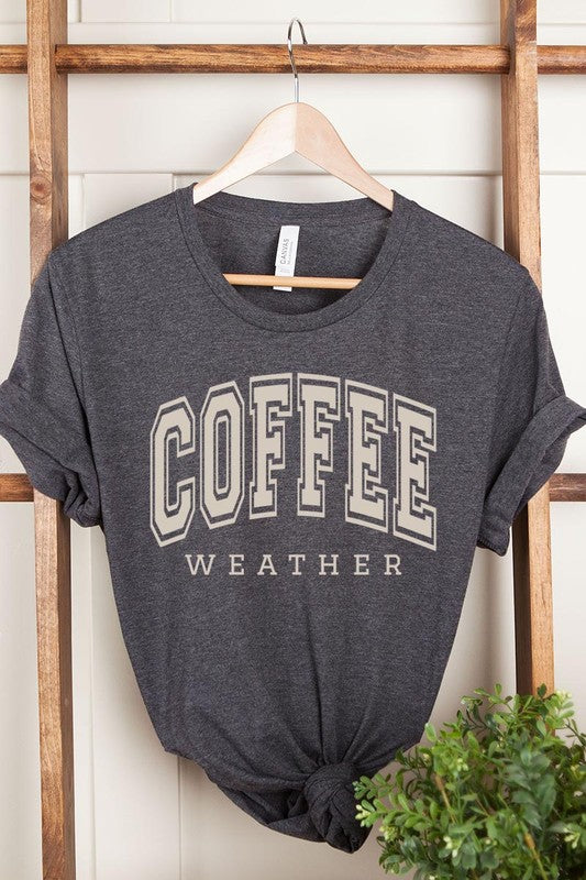 The Coffee Weather Graphic Tee, in a white hue with bold brown text, hangs on a wooden hanger, complemented by a beige sweater draped beside it and set against a background of lush green foliage. This unisex crew neck short sleeve tee showcases an eye-catching graphic design created through high-quality direct-to-film printing.