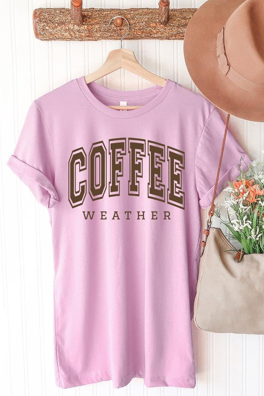 The Coffee Weather Graphic Tee, in a white hue with bold brown text, hangs on a wooden hanger, complemented by a beige sweater draped beside it and set against a background of lush green foliage. This unisex crew neck short sleeve tee showcases an eye-catching graphic design created through high-quality direct-to-film printing.