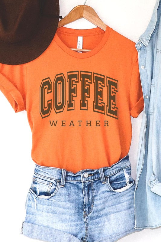 The Coffee Weather Graphic Tee, in a white hue with bold brown text, hangs on a wooden hanger, complemented by a beige sweater draped beside it and set against a background of lush green foliage. This unisex crew neck short sleeve tee showcases an eye-catching graphic design created through high-quality direct-to-film printing.