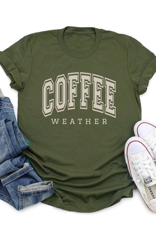 The Coffee Weather Graphic Tee, in a white hue with bold brown text, hangs on a wooden hanger, complemented by a beige sweater draped beside it and set against a background of lush green foliage. This unisex crew neck short sleeve tee showcases an eye-catching graphic design created through high-quality direct-to-film printing.