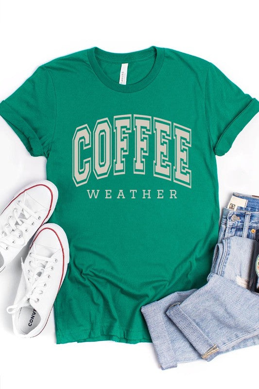 The Coffee Weather Graphic Tee, in a white hue with bold brown text, hangs on a wooden hanger, complemented by a beige sweater draped beside it and set against a background of lush green foliage. This unisex crew neck short sleeve tee showcases an eye-catching graphic design created through high-quality direct-to-film printing.