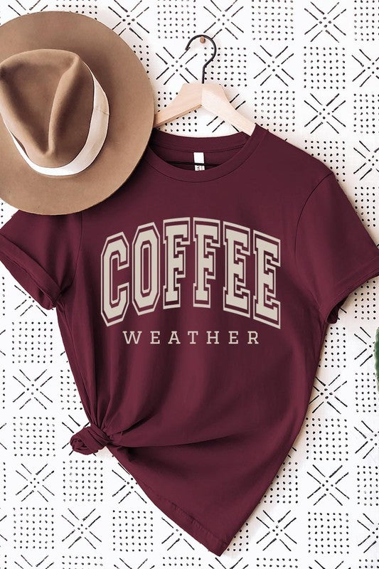 The Coffee Weather Graphic Tee, in a white hue with bold brown text, hangs on a wooden hanger, complemented by a beige sweater draped beside it and set against a background of lush green foliage. This unisex crew neck short sleeve tee showcases an eye-catching graphic design created through high-quality direct-to-film printing.