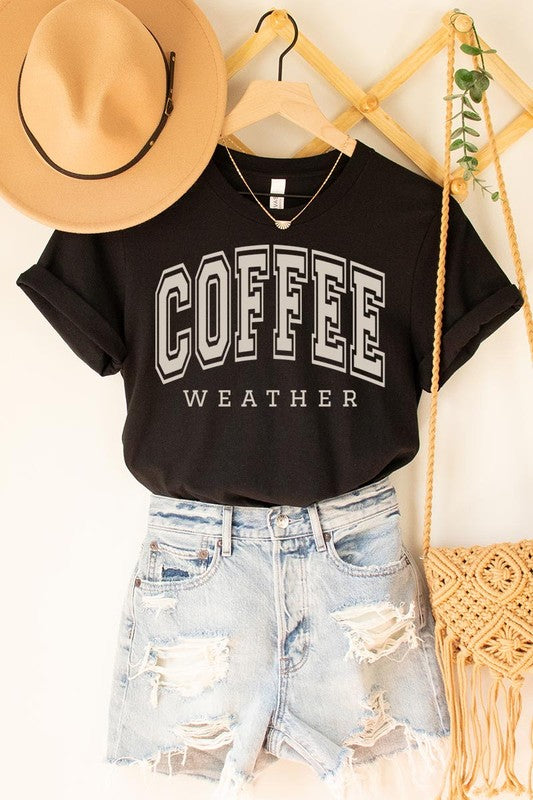 The Coffee Weather Graphic Tee, in a white hue with bold brown text, hangs on a wooden hanger, complemented by a beige sweater draped beside it and set against a background of lush green foliage. This unisex crew neck short sleeve tee showcases an eye-catching graphic design created through high-quality direct-to-film printing.