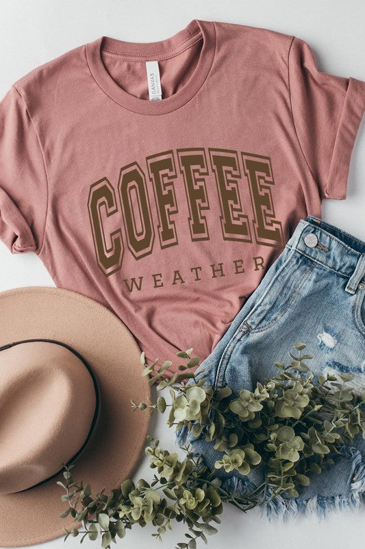 The Coffee Weather Graphic Tee, in a white hue with bold brown text, hangs on a wooden hanger, complemented by a beige sweater draped beside it and set against a background of lush green foliage. This unisex crew neck short sleeve tee showcases an eye-catching graphic design created through high-quality direct-to-film printing.