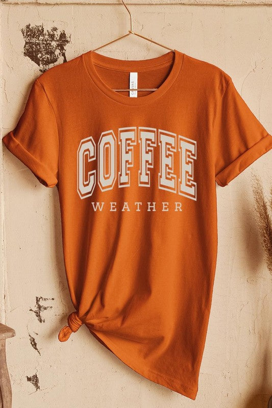 The Coffee Weather Graphic Tee, in a white hue with bold brown text, hangs on a wooden hanger, complemented by a beige sweater draped beside it and set against a background of lush green foliage. This unisex crew neck short sleeve tee showcases an eye-catching graphic design created through high-quality direct-to-film printing.