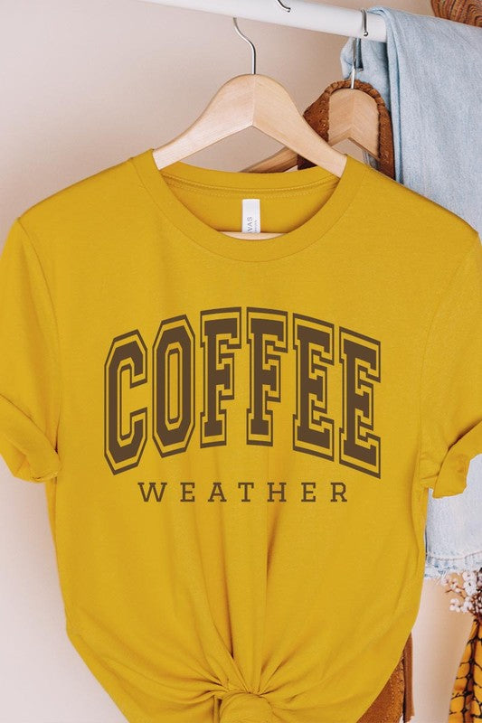 The Coffee Weather Graphic Tee, in a white hue with bold brown text, hangs on a wooden hanger, complemented by a beige sweater draped beside it and set against a background of lush green foliage. This unisex crew neck short sleeve tee showcases an eye-catching graphic design created through high-quality direct-to-film printing.