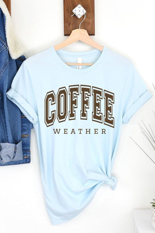 The Coffee Weather Graphic Tee, in a white hue with bold brown text, hangs on a wooden hanger, complemented by a beige sweater draped beside it and set against a background of lush green foliage. This unisex crew neck short sleeve tee showcases an eye-catching graphic design created through high-quality direct-to-film printing.