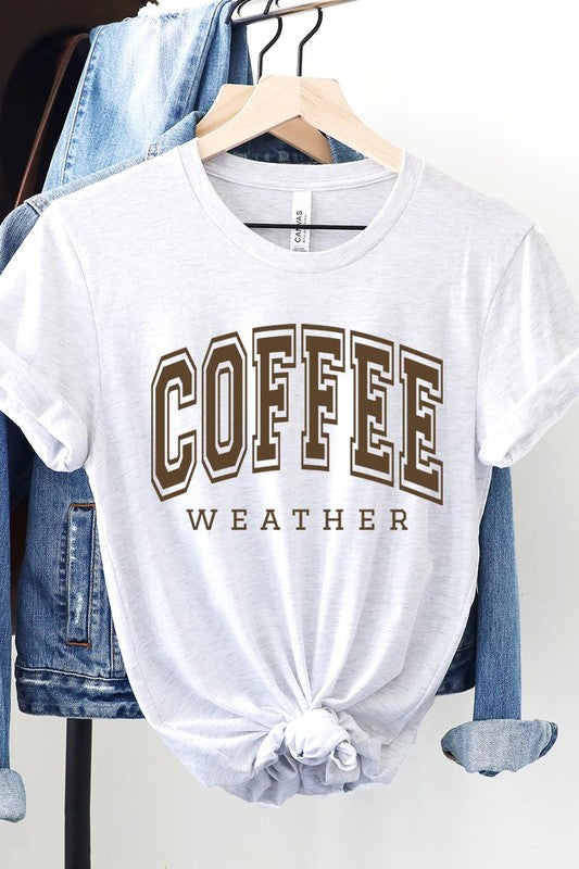 The Coffee Weather Graphic Tee, in a white hue with bold brown text, hangs on a wooden hanger, complemented by a beige sweater draped beside it and set against a background of lush green foliage. This unisex crew neck short sleeve tee showcases an eye-catching graphic design created through high-quality direct-to-film printing.