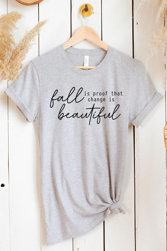 The "Fall Is Proof That Graphic Tee" with rolled sleeves, featuring the printed text "Fall is Proof that change is beautiful," hangs on a wall. This unisex crew neck shirt also sports a small tying knot at the bottom.