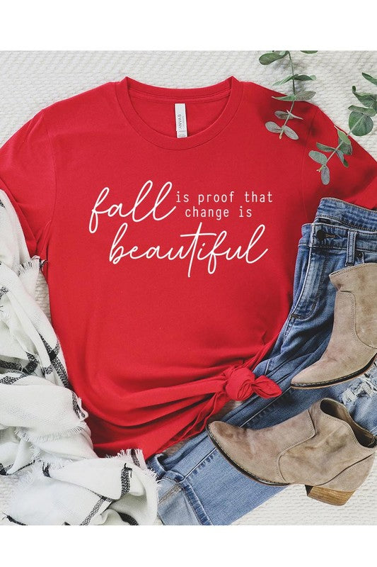 The "Fall Is Proof That Graphic Tee" with rolled sleeves, featuring the printed text "Fall is Proof that change is beautiful," hangs on a wall. This unisex crew neck shirt also sports a small tying knot at the bottom.