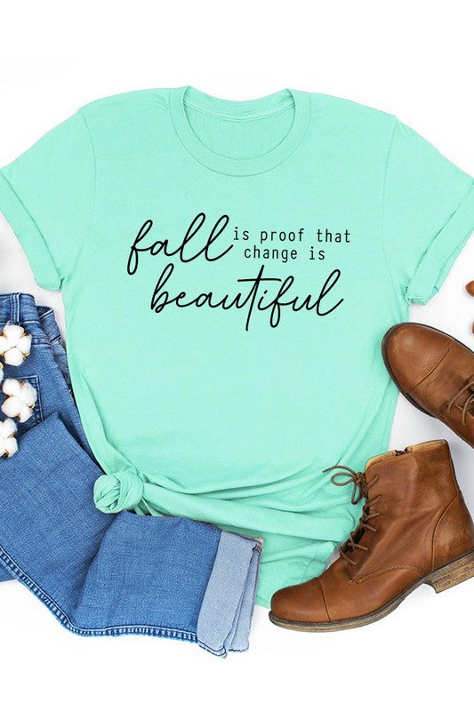 The "Fall Is Proof That Graphic Tee" with rolled sleeves, featuring the printed text "Fall is Proof that change is beautiful," hangs on a wall. This unisex crew neck shirt also sports a small tying knot at the bottom.