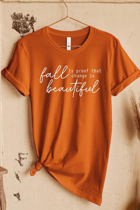 The "Fall Is Proof That Graphic Tee" with rolled sleeves, featuring the printed text "Fall is Proof that change is beautiful," hangs on a wall. This unisex crew neck shirt also sports a small tying knot at the bottom.