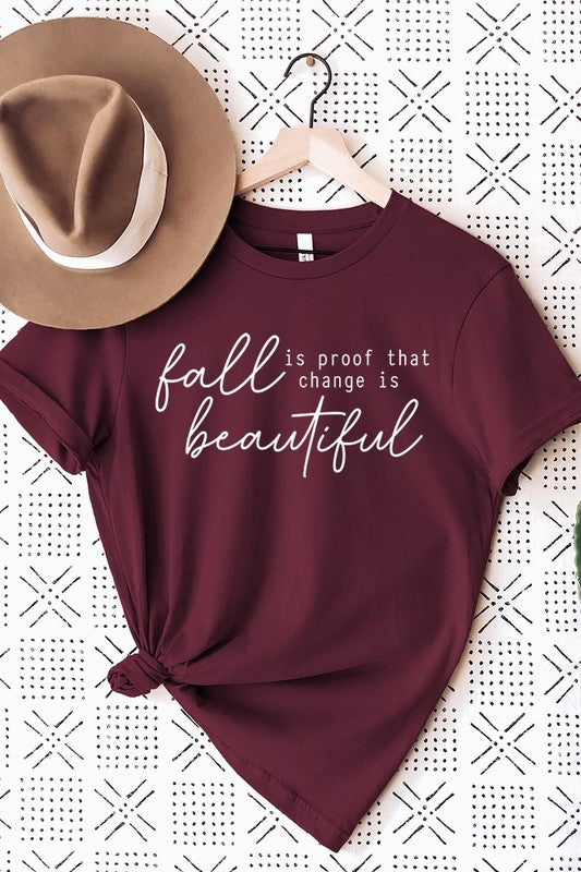 The "Fall Is Proof That Graphic Tee" with rolled sleeves, featuring the printed text "Fall is Proof that change is beautiful," hangs on a wall. This unisex crew neck shirt also sports a small tying knot at the bottom.