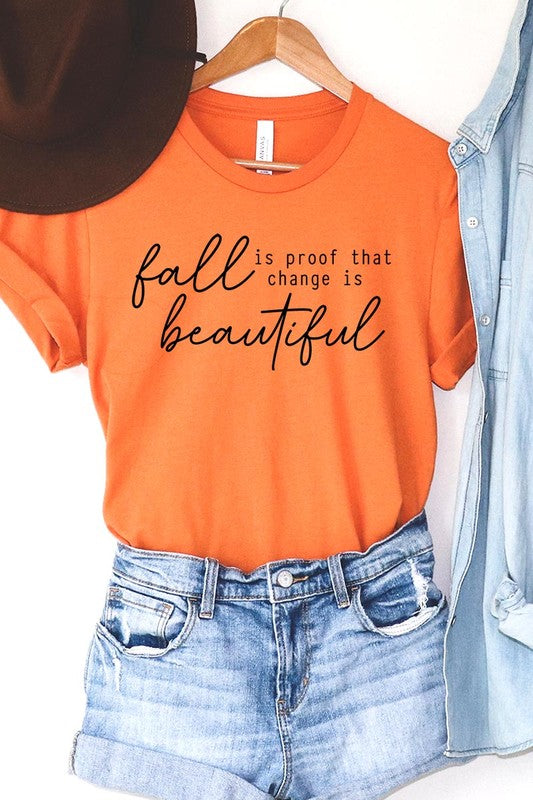 The "Fall Is Proof That Graphic Tee" with rolled sleeves, featuring the printed text "Fall is Proof that change is beautiful," hangs on a wall. This unisex crew neck shirt also sports a small tying knot at the bottom.