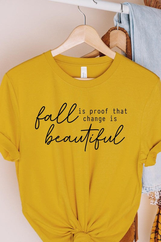 The "Fall Is Proof That Graphic Tee" with rolled sleeves, featuring the printed text "Fall is Proof that change is beautiful," hangs on a wall. This unisex crew neck shirt also sports a small tying knot at the bottom.