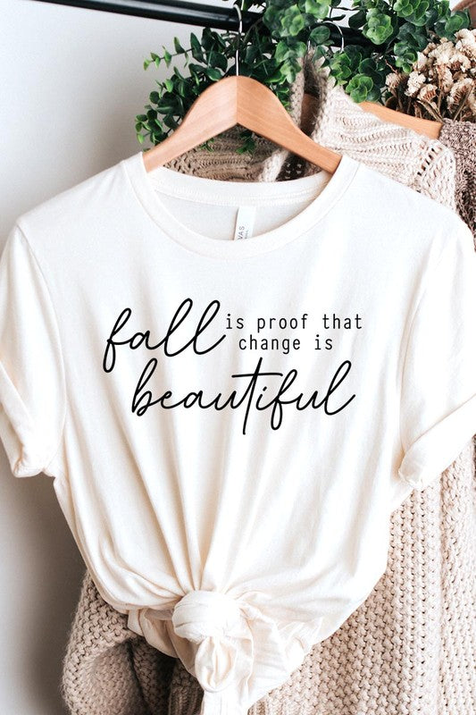 The "Fall Is Proof That Graphic Tee" with rolled sleeves, featuring the printed text "Fall is Proof that change is beautiful," hangs on a wall. This unisex crew neck shirt also sports a small tying knot at the bottom.