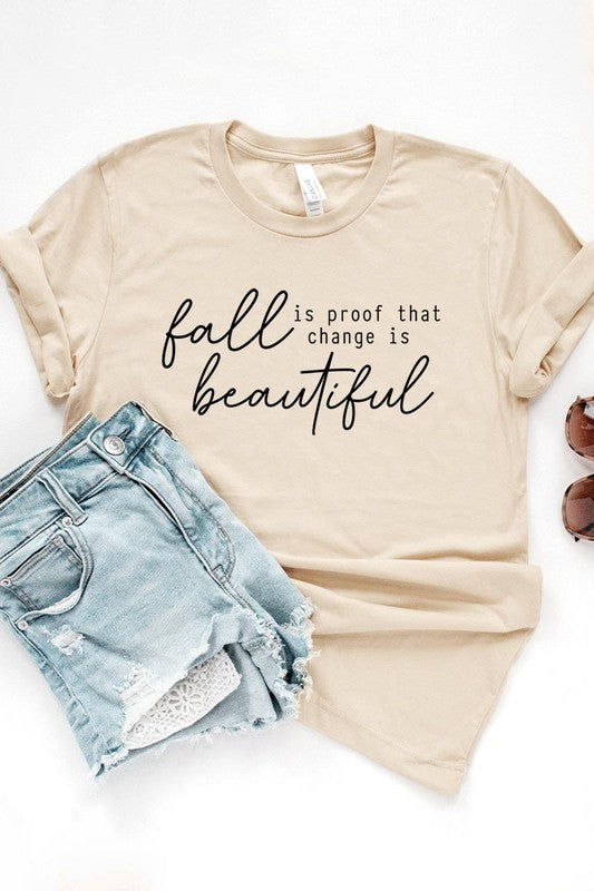 The "Fall Is Proof That Graphic Tee" with rolled sleeves, featuring the printed text "Fall is Proof that change is beautiful," hangs on a wall. This unisex crew neck shirt also sports a small tying knot at the bottom.