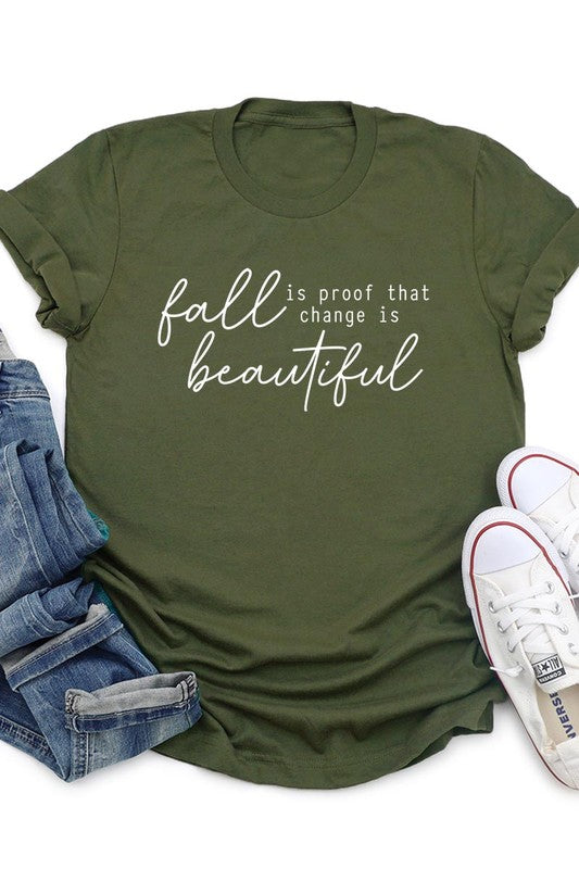 The "Fall Is Proof That Graphic Tee" with rolled sleeves, featuring the printed text "Fall is Proof that change is beautiful," hangs on a wall. This unisex crew neck shirt also sports a small tying knot at the bottom.