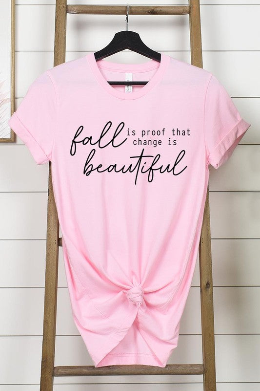 The "Fall Is Proof That Graphic Tee" with rolled sleeves, featuring the printed text "Fall is Proof that change is beautiful," hangs on a wall. This unisex crew neck shirt also sports a small tying knot at the bottom.