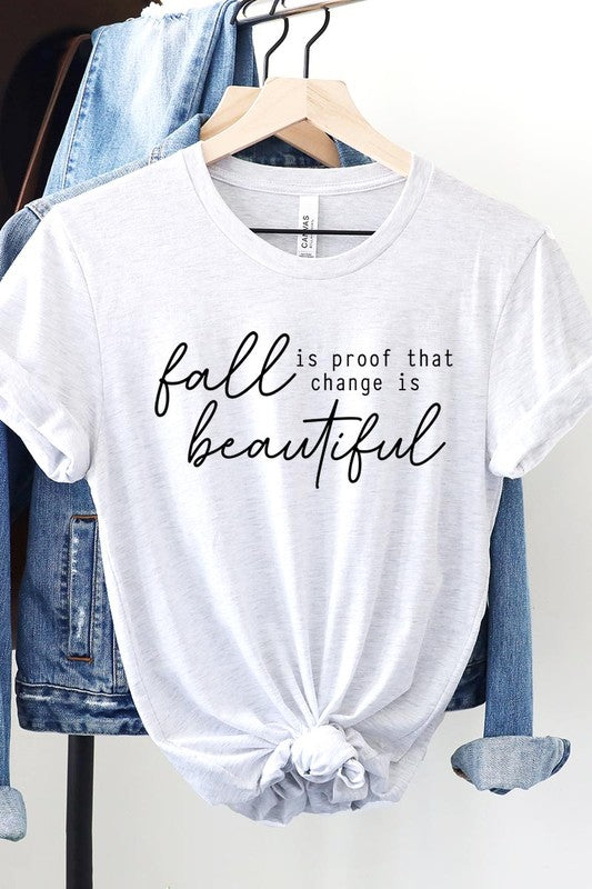 The "Fall Is Proof That Graphic Tee" with rolled sleeves, featuring the printed text "Fall is Proof that change is beautiful," hangs on a wall. This unisex crew neck shirt also sports a small tying knot at the bottom.