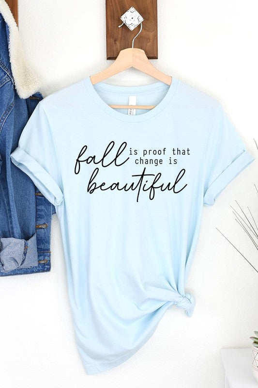 The "Fall Is Proof That Graphic Tee" with rolled sleeves, featuring the printed text "Fall is Proof that change is beautiful," hangs on a wall. This unisex crew neck shirt also sports a small tying knot at the bottom.