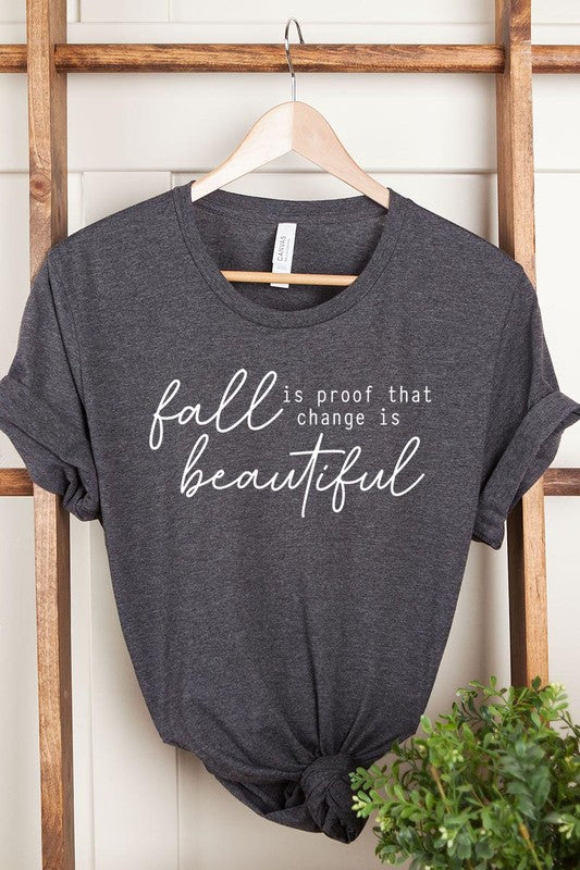 The "Fall Is Proof That Graphic Tee" with rolled sleeves, featuring the printed text "Fall is Proof that change is beautiful," hangs on a wall. This unisex crew neck shirt also sports a small tying knot at the bottom.