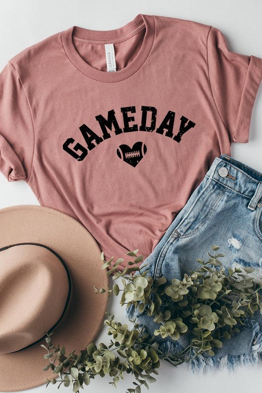 A red Game Day Football Heart Graphic Tee featuring the word "GAMEDAY" with a football inside a heart graphic below it, laid on a white surface paired with blue jeans, a striped scarf, and tan ankle boots. Ideal for Family Group Uniforms on game day!