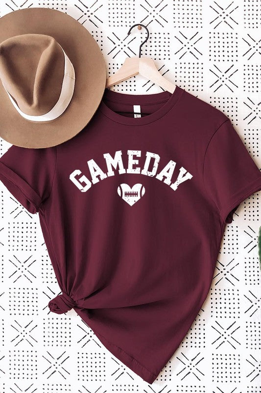 A red Game Day Football Heart Graphic Tee featuring the word "GAMEDAY" with a football inside a heart graphic below it, laid on a white surface paired with blue jeans, a striped scarf, and tan ankle boots. Ideal for Family Group Uniforms on game day!