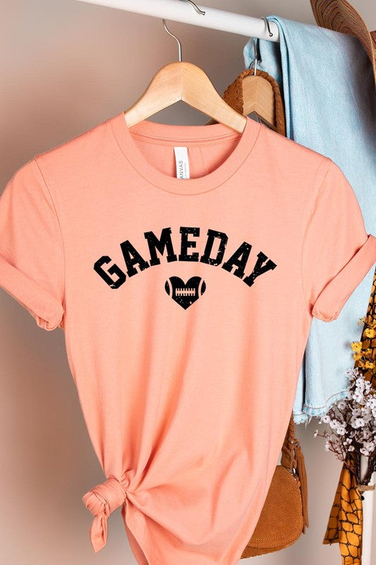 A red Game Day Football Heart Graphic Tee featuring the word "GAMEDAY" with a football inside a heart graphic below it, laid on a white surface paired with blue jeans, a striped scarf, and tan ankle boots. Ideal for Family Group Uniforms on game day!