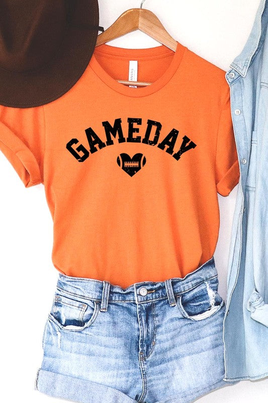 A red Game Day Football Heart Graphic Tee featuring the word "GAMEDAY" with a football inside a heart graphic below it, laid on a white surface paired with blue jeans, a striped scarf, and tan ankle boots. Ideal for Family Group Uniforms on game day!