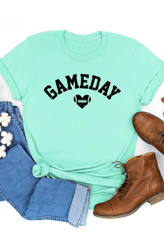 A red Game Day Football Heart Graphic Tee featuring the word "GAMEDAY" with a football inside a heart graphic below it, laid on a white surface paired with blue jeans, a striped scarf, and tan ankle boots. Ideal for Family Group Uniforms on game day!