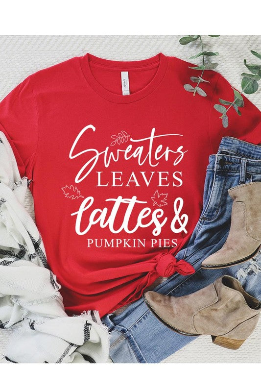 A white "Sweaters,Leaves Graphic Tee" hangs on a wooden hanger, featuring the text "Sweaters, Leaves, Lattes & Pumpkin Pies." In the background, a cozy knitted sweater adds to the inviting scene. This unisex T-shirt blends seasonal charm with everyday comfort.