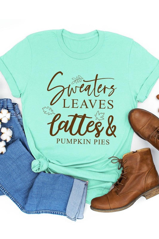 A white "Sweaters,Leaves Graphic Tee" hangs on a wooden hanger, featuring the text "Sweaters, Leaves, Lattes & Pumpkin Pies." In the background, a cozy knitted sweater adds to the inviting scene. This unisex T-shirt blends seasonal charm with everyday comfort.