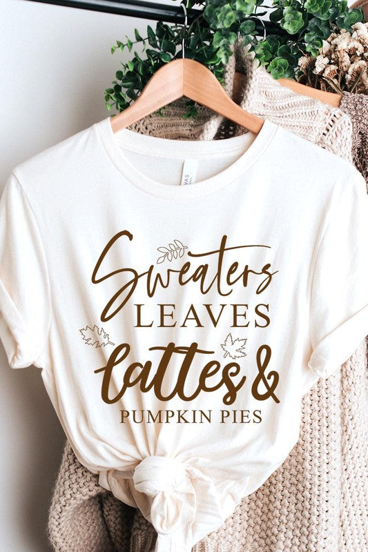 A white "Sweaters,Leaves Graphic Tee" hangs on a wooden hanger, featuring the text "Sweaters, Leaves, Lattes & Pumpkin Pies." In the background, a cozy knitted sweater adds to the inviting scene. This unisex T-shirt blends seasonal charm with everyday comfort.