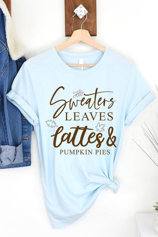 A white "Sweaters,Leaves Graphic Tee" hangs on a wooden hanger, featuring the text "Sweaters, Leaves, Lattes & Pumpkin Pies." In the background, a cozy knitted sweater adds to the inviting scene. This unisex T-shirt blends seasonal charm with everyday comfort.