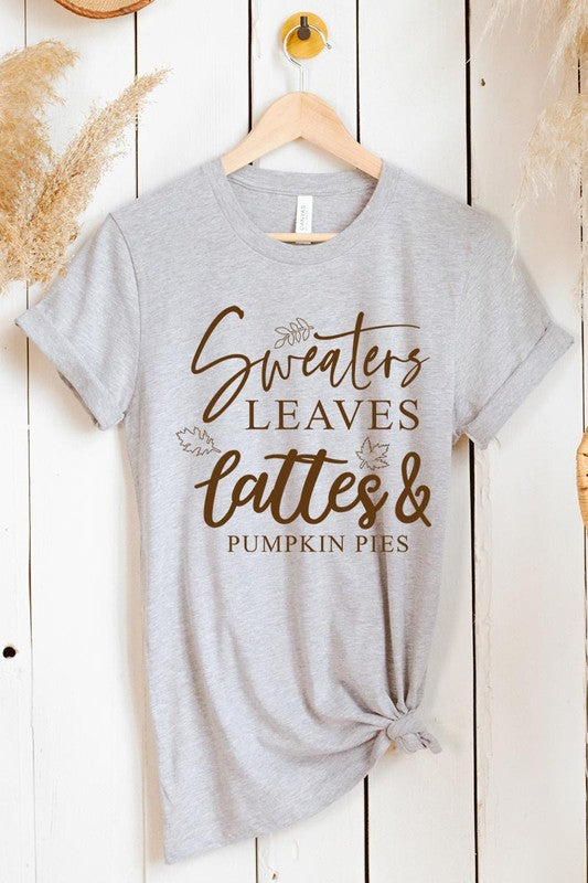 A white "Sweaters,Leaves Graphic Tee" hangs on a wooden hanger, featuring the text "Sweaters, Leaves, Lattes & Pumpkin Pies." In the background, a cozy knitted sweater adds to the inviting scene. This unisex T-shirt blends seasonal charm with everyday comfort.